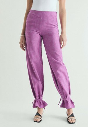Mujer que lleva pantalones lavanda de talle alto con detalles de volantes y lazos en los tobillos, combinados con sandalias negras de tiras y una camiseta blanca sin mangas.