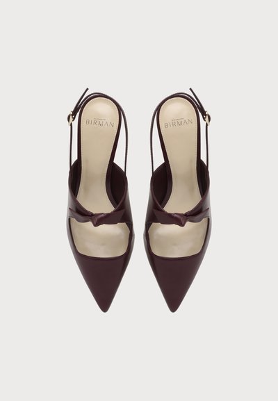 Ζευγάρι μπορντό slingback γόβες με μυτερή μύτη, πλαϊνά λουράκια με αγκράφες και διακοσμητικούς φιόγκους μπροστά, φωτογραφημένα από πάνω σε λευκό φόντο.