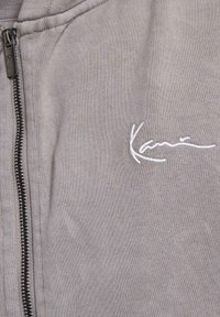 Prenda de color gris claro con cremalleras negras y bordado en cursiva blanca que dice "Kane" en el lado frontal.