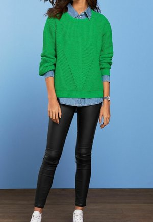 Femme portant un pull vert en maille sur une chemise en denim bleu, un pantalon en cuir noir, des baskets blanches et une montre-bracelet argentée, debout devant un fond bleu.