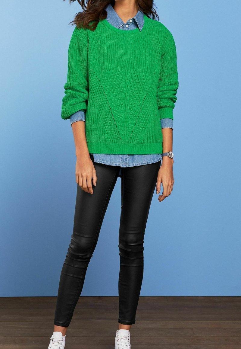 Vrouw die een groene gebreide trui draagt over een blauw denim overhemd, zwarte leren broek, witte sneakers en een zilveren polshorloge, staand tegen een blauwe achtergrond.