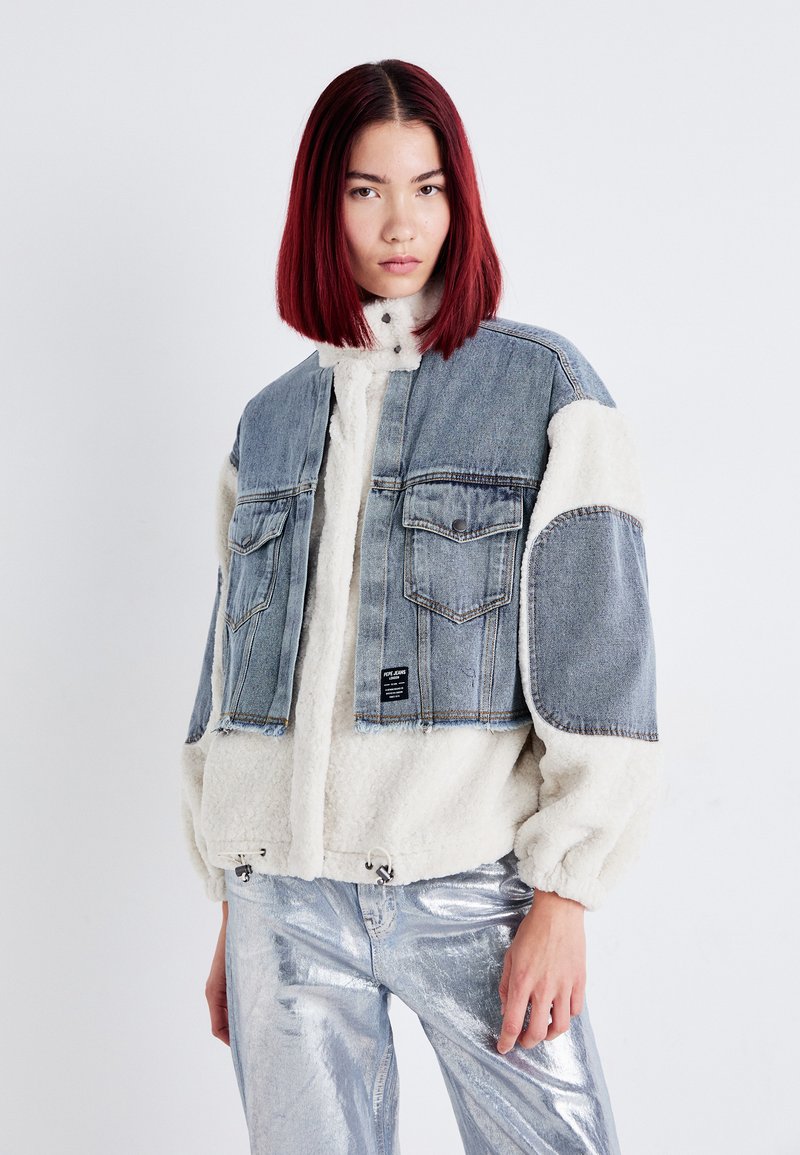 Denim- og faux fur cropped jakke med blå denim overdel, hvide fluffy ærmer, to brystlommer og snoretræk i kanten. Sølvskinnende bukser.