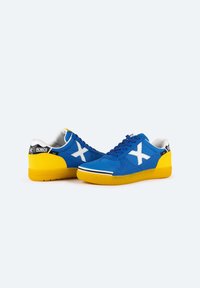 Zapatillas deportivas azules con acentos blancos, presentan un upper texturizado, suela de goma amarilla y un distintivo logo X blanco en los laterales.