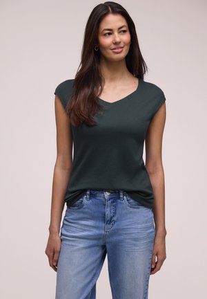 Femme aux longs cheveux foncés portant un haut à manches courtes vert foncé et un jean bleu clair, debout devant un fond clair uni.