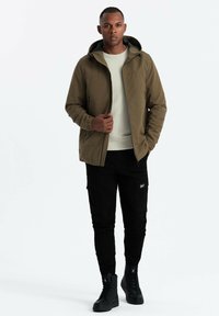 Giacca khaki con zip e cappuccio, tessuto testurizzato, abbinata a pantaloni cargo neri e stivali alti neri. Design semplice e funzionale.