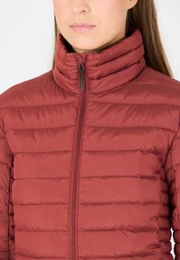 Rød polstret jakke med høj krave og lodrette syninger. Har en frontlynlås med sort hardware og en glat, blank tekstur.