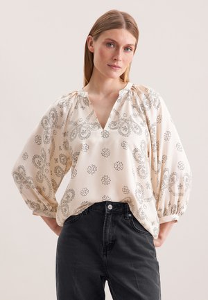 Blusa crema con maniche a palloncino, scollo a V e motivi floreali neri. Abbinata a jeans neri a vita alta. Texture del tessuto liscia.