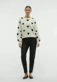 Pulover alb cu model de inimi negre, guler rotund și mâneci lungi, asortat cu pantaloni negri și pantofi negri. Textură netedă.