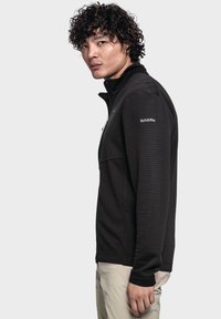 Schwarze Fleecejacke mit Reißverschluss und strukturiertem Rippmuster, mit hohem Kragen und Logo auf der Schulter. Tragen Sie sie mit hellen Hosen.