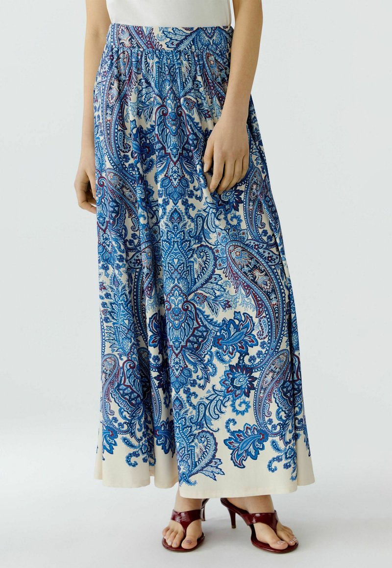 Langer Rock für Damen mit blauen Paisley-Blumenmustern auf weißem Stoff, kombiniert mit braunen Sandalen mit Absatz und weißem Oberteil, stehend vor einfachem Hintergrund.