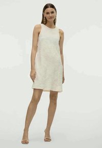 Vestido A-line em off-white, sem mangas, com um padrão floral subtil, até ao joelho, combinado com sandálias de salto abertas em bege.