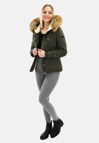Young woman smiling, wearing a green fur-hooded jacket, black top, gray skinny jeans, and black ankle boots, standing on a plain background.