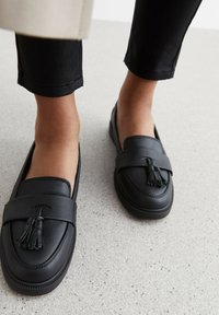 Mocassini in pelle nera con un accento a nappina, caratterizzati da una punta tonda e dettagli cuciti, indossati con pantaloni neri su una superficie grigio chiaro.