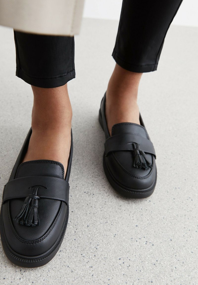 Mocassini in pelle nera con un accento a nappina, caratterizzati da una punta tonda e dettagli cuciti, indossati con pantaloni neri su una superficie grigio chiaro.