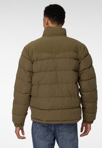 Olivenfarget puffjakke med høy krage, ribbede detaljer og quiltet tekstur. Baksiden har horisontal søm og elastiske mansjetter.