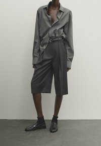 Chemise slim-fit grise à rayures avec un col ouvert, associée à un short gris taille haute plissé. Complétée par des chaussures noires et des chaussettes.