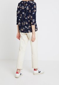 Navyblaues Blumenbluse mit Dreiviertelärmeln, kombiniert mit hellbeigen, knöchellangen Hosen und weißen Turnschuhen mit roten und schwarzen Akzenten.