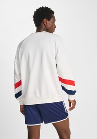 Man in een witte sweatshirt met rood en marineblauwe strepen op de mouwen en marineblauwe sportshorts, staande met zijn rug naar de camera tegen een plain witte achtergrond.