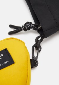 Petite pochette jaune à surface texturée, fixée à un sac en tissu noir par une boucle et un mousqueton noir ; étiquette de marque noire.