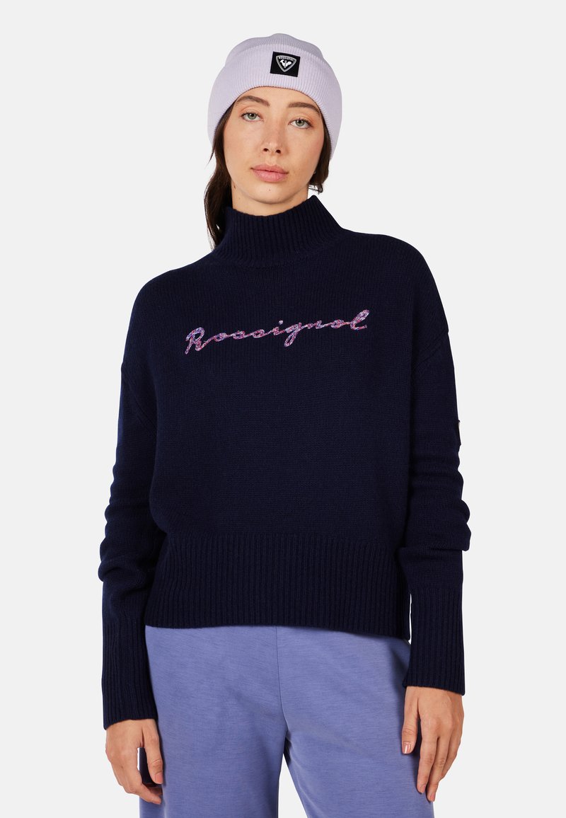 Navy knitted turtleneck sweater featuring a ribbed hem and cuffs, with pink script logo "Rossignol" across the front. Paired with light blue pants.