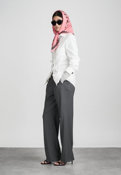 Witte geraffineerde blouse, grijze wijde broek, roze geblokte sjaal en zwarte zonnebril. De sandalen hebben dunne bandjes en een hoge hak.