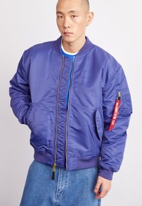 Chaqueta bomber morada hecha de nylon suave, con cierre frontal, puños acanalados y un bolsillo con parche de acento rojo en la manga.