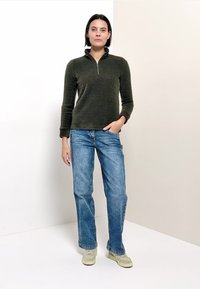 Grüner Pullover mit Reißverschluss, blaue Jeans und helle Sneakers. Der Pullover hat einen hohen Kragen und eine figurbetonte Passform; die Jeans sind mit geradem Bein und klassischem Waschung.