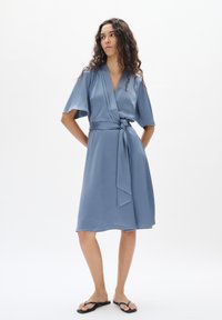 Knielanges Wickelkleid aus weichem blauen Stoff mit texturierter Oberfläche. Verfügt über kurze Ärmel, eine gebundene Taille und eine lockere Passform. Kombiniert mit schwarzen Flip-Flops.