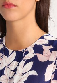 Marinblus med blommönster, prydd med stora ljusrosa blad. Slät textur, rund halsringning och subtil glans i tyget.