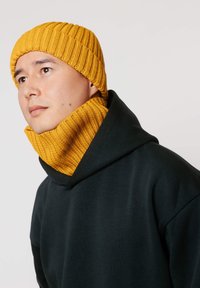 Żółta prążkowana dzianinowa czapka i pasująca do niej komin z golfem, noszone na ciemnozielonym kapturze. Charakteryzują się teksturowanymi wzorami dzianiny i luźnym krojem.