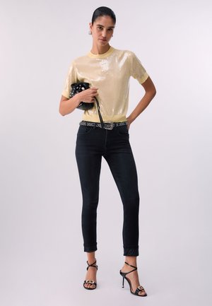 Femme portant un haut à manches courtes orné de sequins jaune pâle, ceinture noire cloutée, jean skinny foncé, sandales à talons hauts noires, tenant un sac noir clouté.