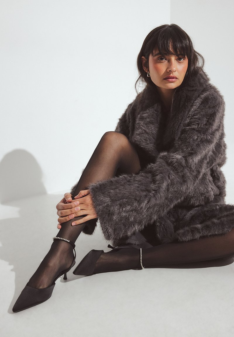 Pelēka mākslīgās kažokādas mētelis ar platiem piedurknēm, uzvilkts pār melnām caurspīdīgām zeķubiksēm un melnām kurpēm ar smailiem purngaliem. Uz kurpēm ir ievērojami potīšu akcenti.