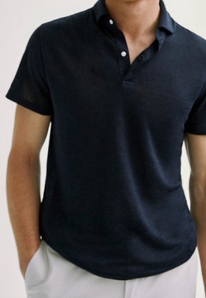 Homme portant un polo noir à manches courtes et un pantalon clair, les mains dans les poches, sur un fond uni.