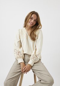 Blusa crema con dettagli in pizzo sulle maniche e sul colletto, caratterizzata da maniche lunghe a sbuffo e design a scollo a V, abbinata a pantaloni a righe beige chiaro.