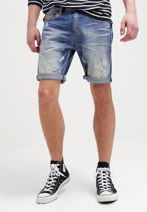 Mężczyzna noszący podwinięte, znoszone szorty z denimu, czarne sneakersy typu high-top z białymi sznurowadłami oraz czarno-białą koszulę w paski.