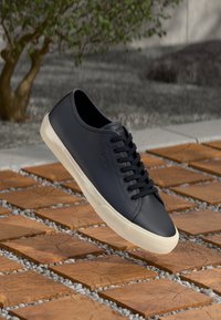 Basket en cuir noir avec semelle en caoutchouc blanche flottant au-dessus de carreaux de bois à motifs et de petits cailloux blancs en extérieur.