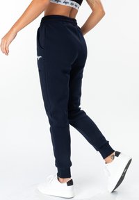 Navy blue sweatpants with elastic waistband and ankle cuffs, featuring side pockets and a white logo accent. Worn with white sneakers.