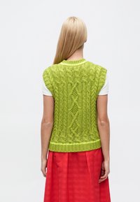 Zaļš adīts bezroku džemperis ar kabeļu rakstiem un apaļu kakla izgriezumu, kopā ar sarkanu svītrainu svārku.