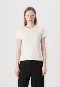 T-shirt di cotone color crema con maniche corte, scollo rotondo e piccolo logo sul petto. Abbinata a pantaloni neri su misura.