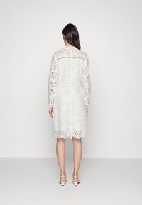 Bílé krajkové šaty s dlouhými rukávy, složitými květinovými vzory, zeleně zakončeným lemem a úzkým pasem, doplněné bílými sandály na vysokém podpatku.