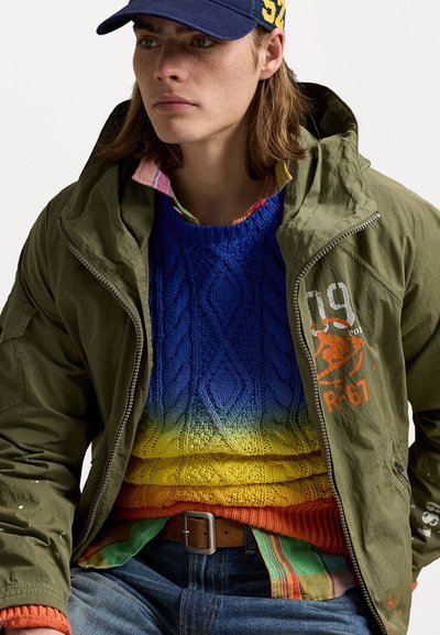 Oliven grøn lynlåsjakke over en blå og gul gradient strikketrøje, med en ternet multikolor skjorte, der stikker ud. Brunt bælte og denim jeans.