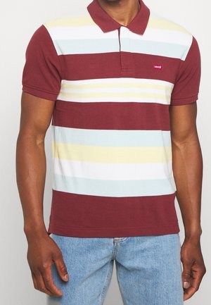 Homme portant un polo à manches courtes avec des rayures horizontales bordeaux, blanc, bleu clair et jaune, associé à un jean bleu clair.