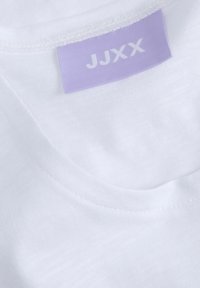Camiseta de algodón blanca con cuello redondo. Presenta una etiqueta lavanda cosida en la tela, que dice "JJXX". Textura suave, diseño minimalista.