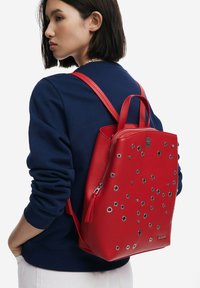 Roter Leder-Rucksack mit runden silbernen Akzenten. Verfügt über einen Reißverschluss, verstellbare Träger und eine strukturierte Form.