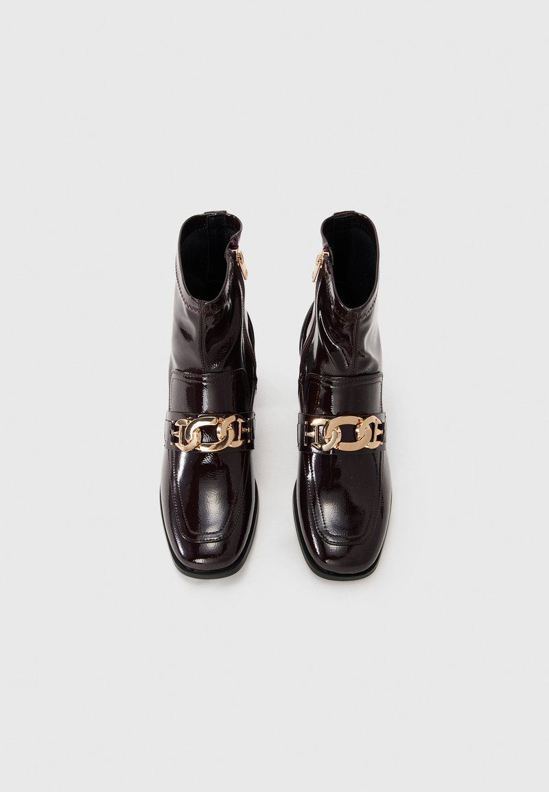 Stivali ankle in pelle verniciata borgogna con punta arrotondata, dettaglio in catena dorata e chiusura con zip sul lato esterno. Texture liscia.