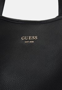 Czarna skórzana torba o teksturowanej powierzchni, z złotym logo "GUESS EST 1981". Uchwyt jest krótki i solidny.