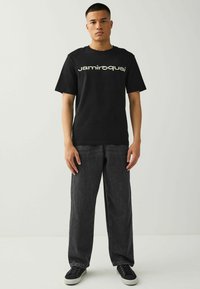 T-shirt di cotone nera con il testo "jamiroquai" in un carattere bianco curvo. Abbinata a pantaloni in denim grigio scuro, larghi, e sneakers nere.