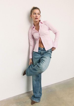 Donna appoggiata a un muro bianco, che indossa una camicia rosa chiaro con bottoni, jeans blu larghi e scarpe marroni con punta, che guarda di lato.