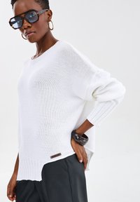Biały, oversize, dzianinowy sweter z prążkowanym dołem, długimi rękawami i bocznym rozcięciem. Posiada metkę z logo na dole. Matowa czarna bransoletka noszona na nadgarstku.