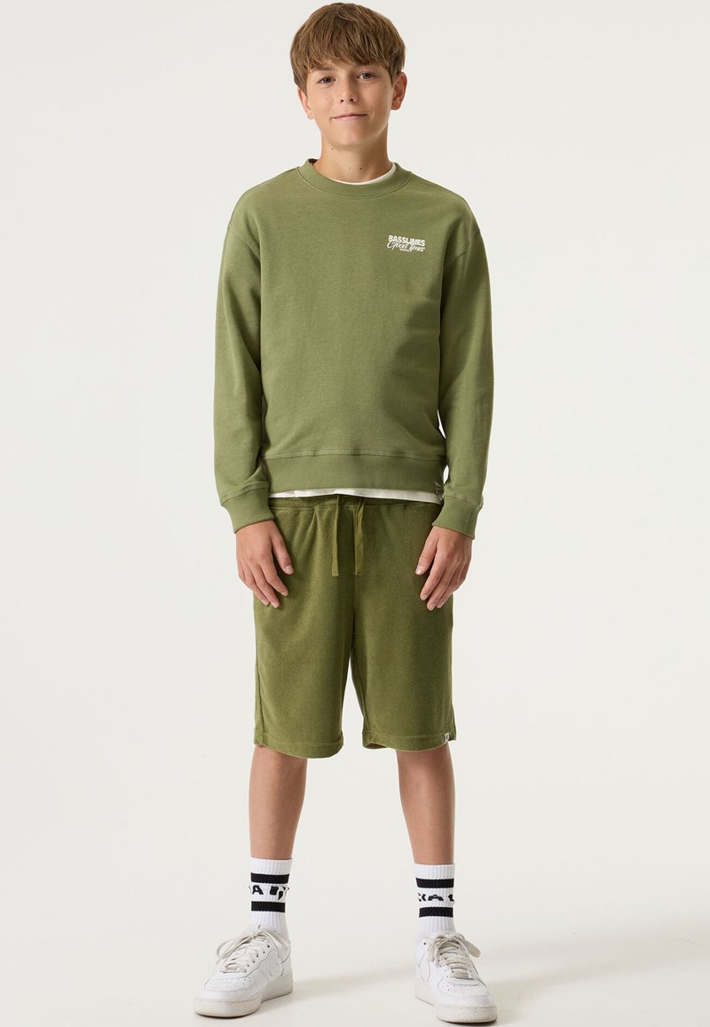 Sweat-shirt vert olive avec poignets et ourlet inférieur côtelés, associé à un short vert olive. Baskets blanches et chaussettes rayées noir et blanc.